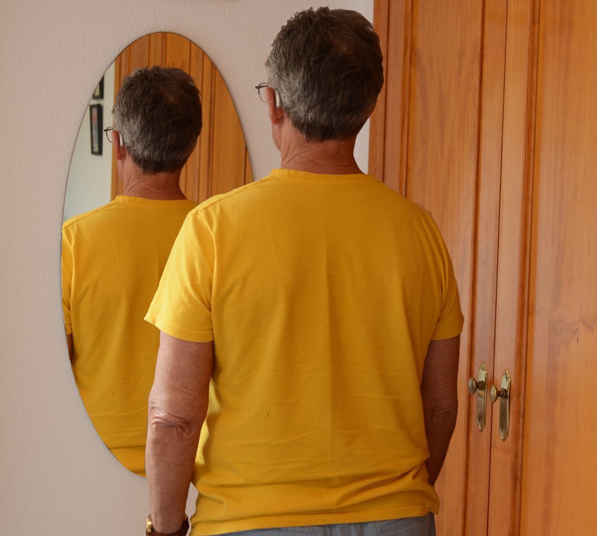 An man looks into a mirror, but instead of seeing his own natural reflection, sees his own back as if caught by a camera capturing him from behind.